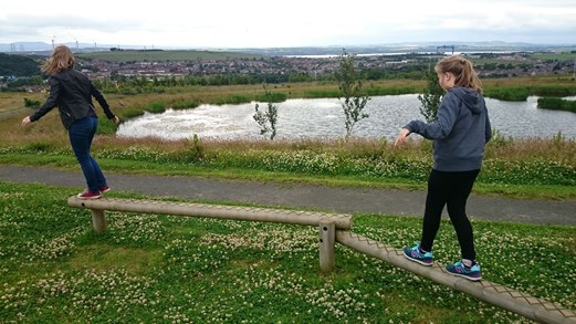 Children balancing on narrow beam at playground. In background, large pond.